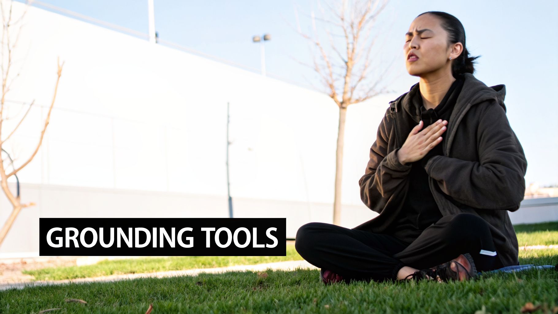A person sits cross-legged on green grass, eyes closed, with hands over their chest, practicing grounding.
