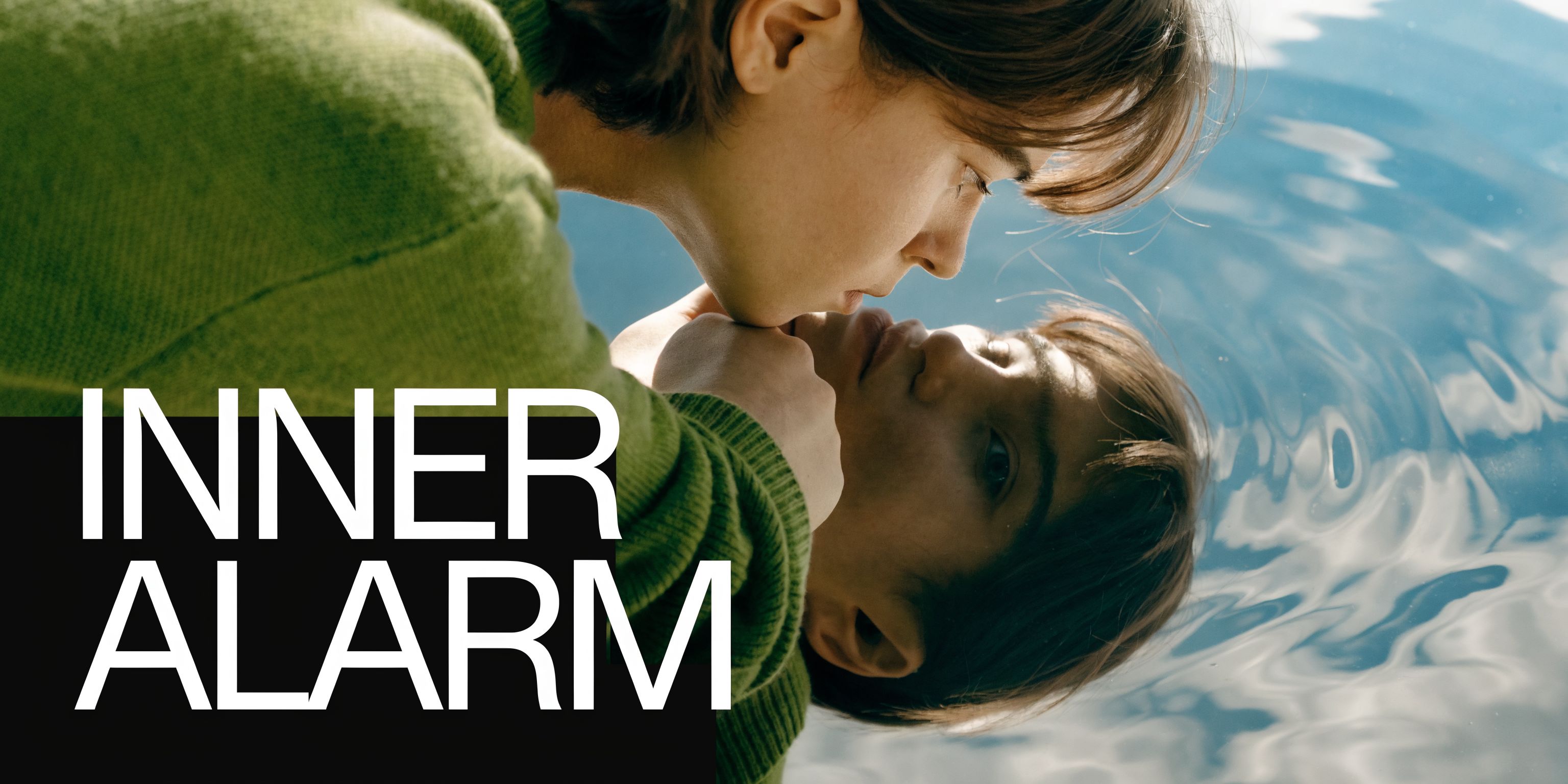 A person in a green sweater looks at their own reflection in the calm blue water surface.