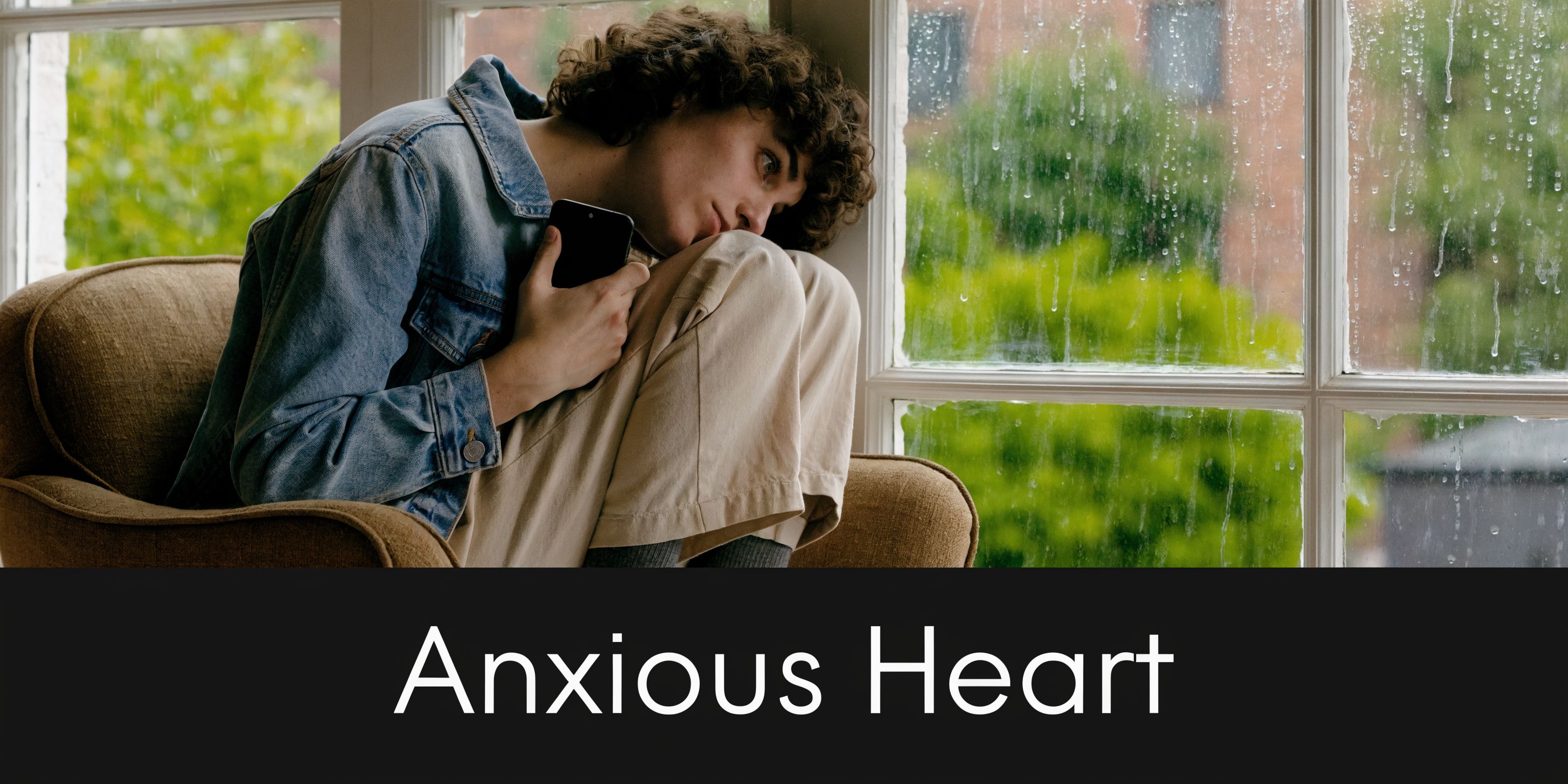 A person with curly hair sitting in an armchair by a rainy window holding a smartphone