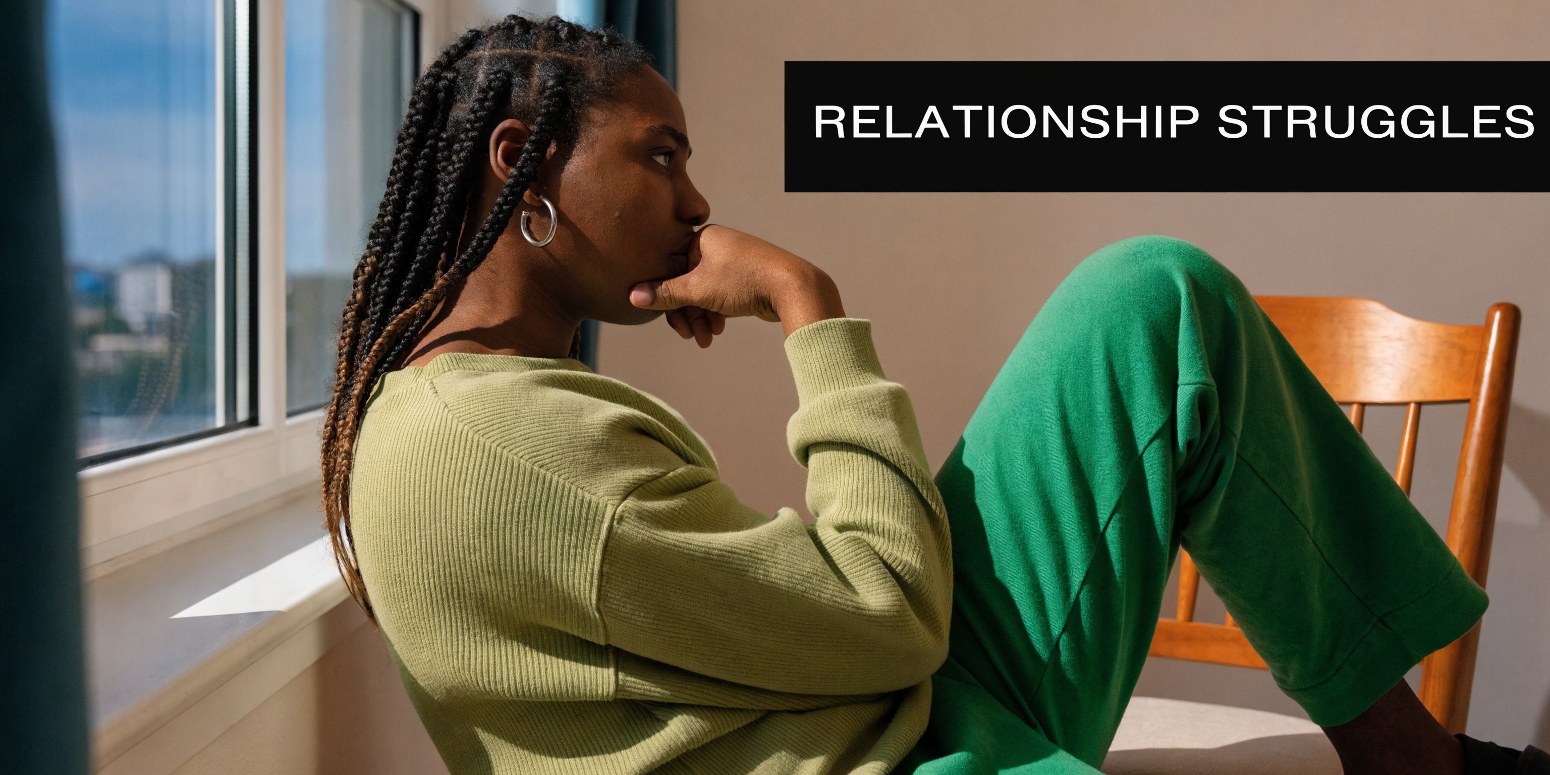 A contemplative young Black woman with braided hair sitting by a window looking away in thought.
