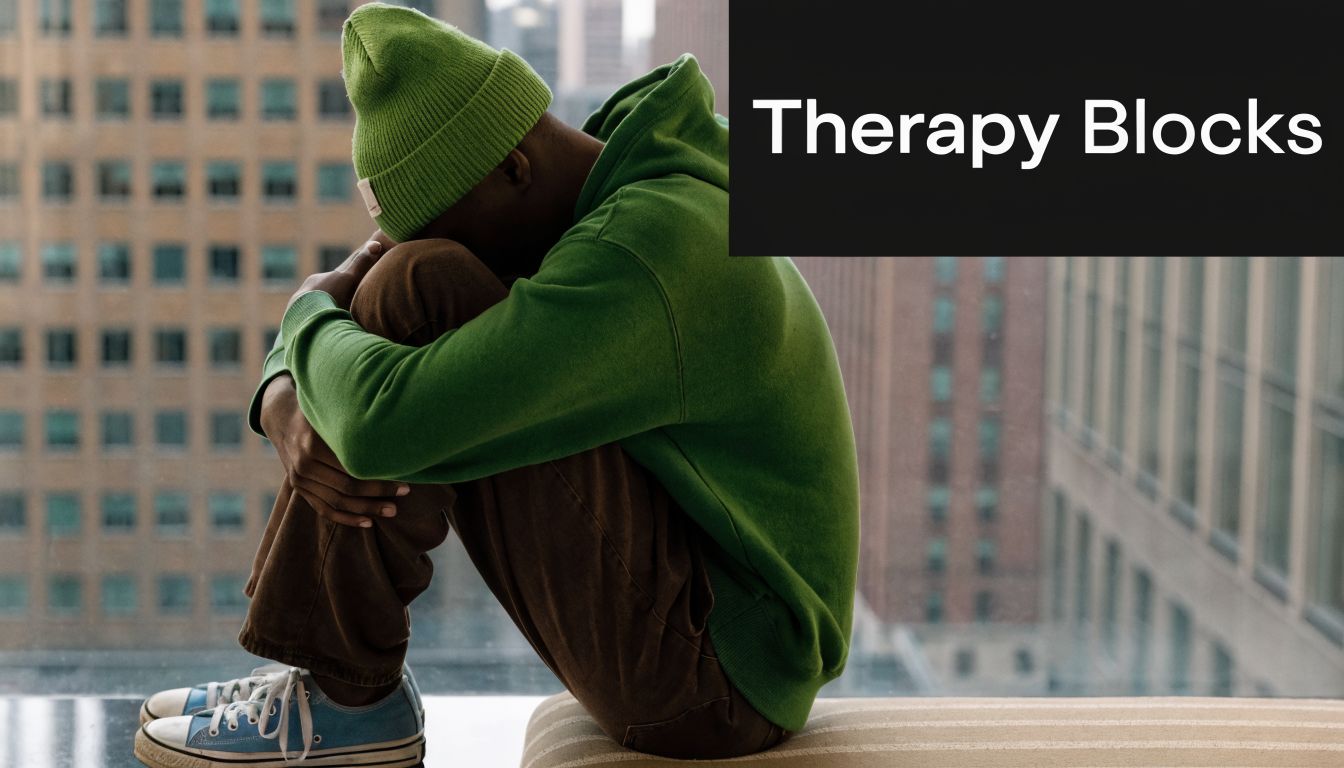 A person wearing a green beanie and hoodie sits curled up looking sad against a city background.